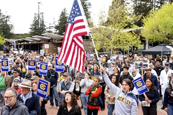 caption: Undergraduate student River Richart hold a flag while protesting against the Trump administration as part of a Day of Action for Higher Education on Thursday, April 17, 2025, in Berkeley, Calif.