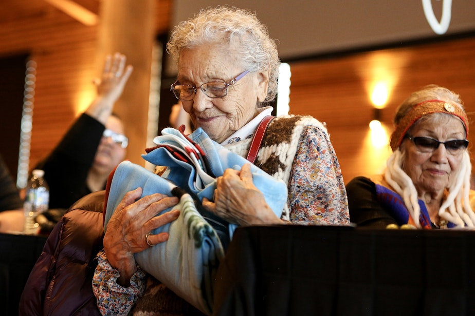 caption: Lummi elder Ernestine Lane receives a gift after sharing her experiences at the Chemawa Indian School in Salem, Oregon. Interior Secretary Deb Haaland visited the Tulalip Gathering Hall on Sunday, April 23, 2023, during the “Road to Healing” tour. The yearlong tour provides opportunity for Indigenous survivors of the federal Indian boarding school system to share their experiences.