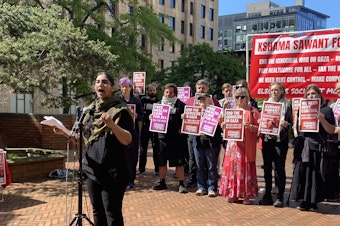 caption: Former Seattle City Councilmember Kshama Sawant stands speaks to supporters outside the Henry M. Jackson Building in downtown Seattle on Monday, June 2. 