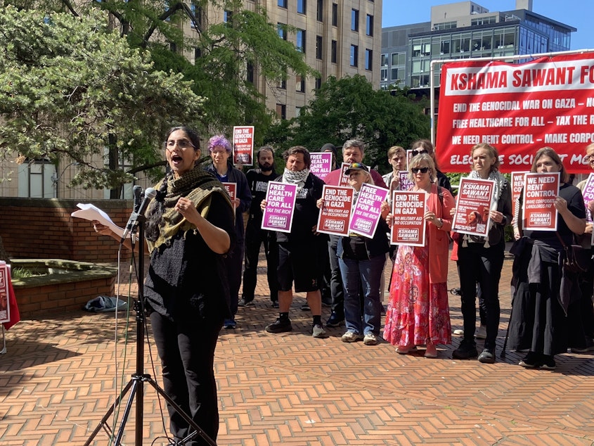 caption: Former Seattle City Councilmember Kshama Sawant stands speaks to supporters outside the Henry M. Jackson Building in downtown Seattle on Monday, June 2.