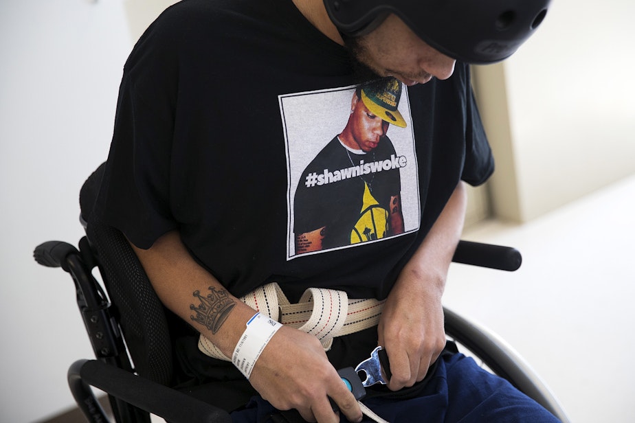 caption: DaShawn Horne buckles the belt of his wheelchair while wearing a t-shirt with his picture and the words #shawniswoke, on Thursday, April 19, 2018, during physical therapy at Harborview Medical Center in Seattle.