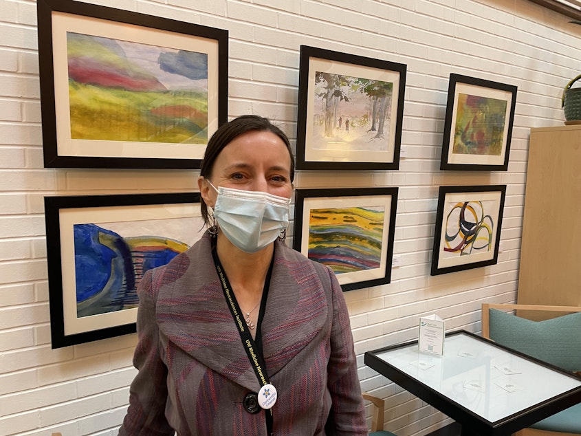 caption: Director of the Memory Hub, Marygrace Becker, stands in front of a wall of watercolor paintings