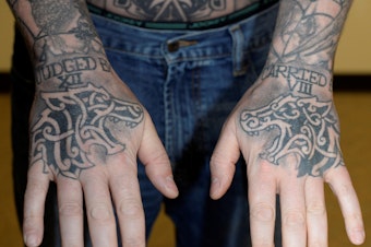 caption: Auburn police officer Jeffrey Nelson displays tattoos on his hands: “JUDGED BY XII” on the right hand. “CARRIED BY VIII” on the left. That’s shorthand for an old policing proverb: “I’d rather be judged by 12” jurors than “carried by six” — or sometimes eight — pallbearers.