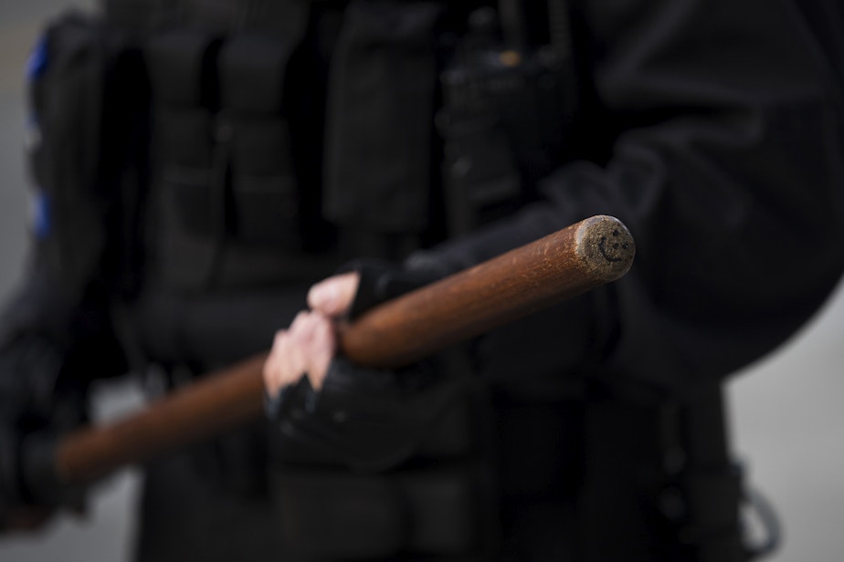 caption: A smiley face is shown drawn on the end of Seattle Police Officer M. Lancaster's baton while standing in a police line on Monday, June 1, 2020, at the intersection of 11th Avenue and East Pine Street in Seattle. For the next week, tense, nightly standoffs between demonstrators and a fortified line of police in riot gear took place at this intersection. On June 8, Seattle police abandoned the East Precinct.