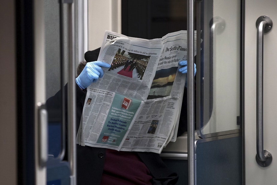 caption: A light rail passenger reads the Seattle Times while wearing blue latex gloves on Thursday, March 26, 2020, at Westlake Station in Seattle.