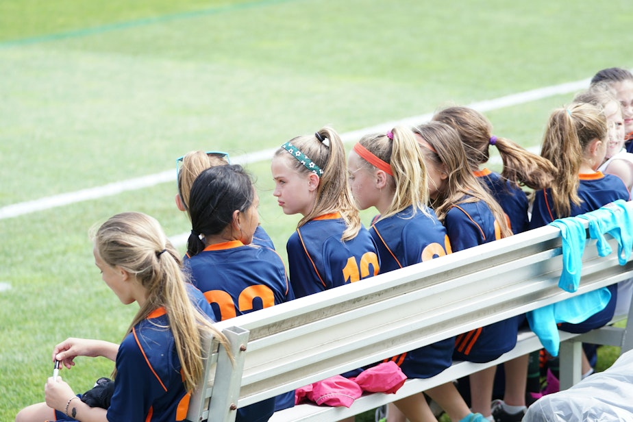caption: A girls' soccer team sits on a bench on the sideline.