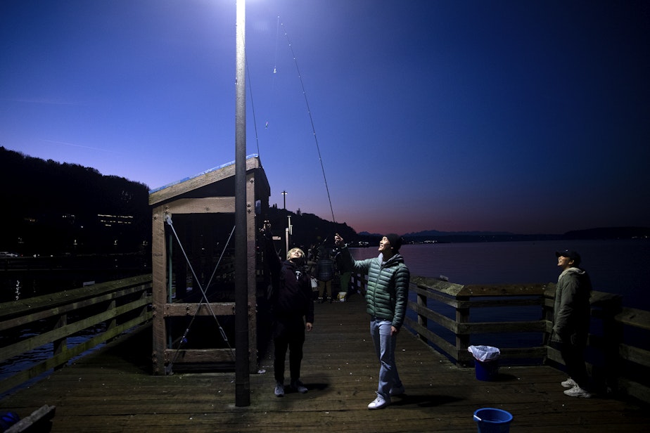 caption: From left, Max, Kody and Deanna, of Denver, hold glow in the dark jigs toward a light before squidding on Friday, November 17, 2023, from Les Davis Pier in Tacoma.